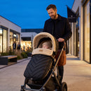 A man pushes a black stroller with a cream fleece-lined hood and quilted chevron footmuff outdoors in a shopping plaza at dusk. A child is sleeping in the stroller.