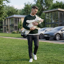 A dad in a green sweater and dark pants walks on a grassy lawn while carrying a baby wearing the Star Fleece Baby Wrap Blanket, with trees, modern houses, and parked cars in the background. | Cosy Baby Blanket - Clair de Lune UK
