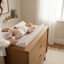 Mum tending to baby lying on the Brown Gingham Anti-Roll Wedge Baby Changing Mat on an oak changing unit but a bright window | Baby Bathing & Changing Essentials - Clair de Lune UK
