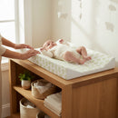 Baby lying on the Leaf Anti-Roll Wedge Baby Changing Mat on an Oak changing unit by the window in a white nursery | Baby Bath Time Essentials - Clair de Lune UK