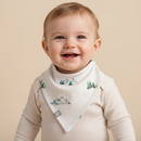 A smiling baby wearing a cream long-sleeve bodysuit and a GrowGrows Little Summit Bib with a green and blue mountain and pine tree pattern, set against a solid beige background.