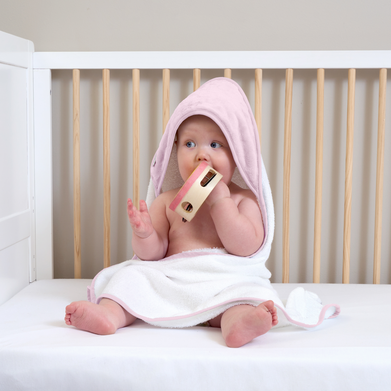 Cute baby sitting in the Essentials Cot Bed Wearing the Pink Marshmallow Hooded Towel and playing with a tambourine | Baby Bathing & Changing Essentials - Clair de Lune UK