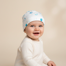 A smiling baby wearing a GrowGrows Marvellous Moons Baby Hat with blue crescent moon and star patterns, paired with a cream long-sleeve bodysuit, set against a warm beige studio background. | Bamboo Babywear - Clair de Lune UK