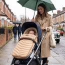 A woman in a beige trench coat pushes a stroller on a city sidewalk while holding a green umbrella. A baby is seated inside the stroller, which is covered with a the beige snug footmuff | Baby Autumn Essentials - Clair de Lune UK