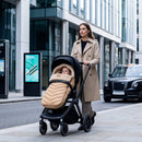 A mum pushes a black stroller with a baby inside on a city sidewalk. The stroller is covered with the beige snug footmuff | Cosytoes | Baby Autumn Essentials - Clair de Lune UK