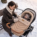 A mother in a winter coat and scarf sits beside a stroller outdoors in the snow, adjusting a beige Snug Universal Pushchair Footmuff with a baby inside. A snow-covered wooden bench is behind them. | Cosytoes - Clair de Lune UK