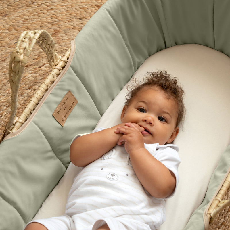 Baby boy smiling in the Sage Green Organic Palm Moses Basket on a natural brown rug | Moses Baskets | Co-sleepers | Nursery Furniture - Clair de Lune UK