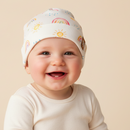 A smiling baby wearing a GrowGrows Radiant Rainbows Baby Hat with a colourful sun and rainbow pattern, paired with a matching cream long-sleeve top, set against a solid beige background. | Bamboo Babywear - Clair de Lune UK