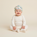 A smiling baby sitting cross-legged on a warm cream studio background, wearing an off-white long-sleeve bodysuit and matching foot socks, and GrowGrows Safari Slumber Baby Hat | Bamboo Babywear - Clair de Lune UK