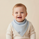 A smiling baby wearing the GrowGrows Sky Blue Baby Bib over a cream long-sleeve top, photographed against a soft beige studio background. | Feeding Essentials - Clair de Lune UK