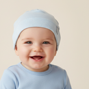 A close-up of a smiling baby wearing the GrowGrows Sky Blue Pinstripe Baby Hat | beanie and a matching light blue long-sleeve top against a soft beige background.
