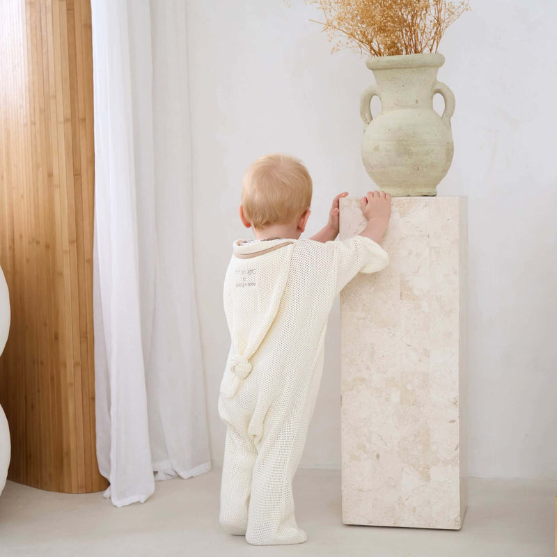 Baby Wearing the Cream Organic Star Breathable Mesh Baby Wrap Blanket standing next to a pedestal with stylish vase arrangement | Cosy Baby Blankets and Swaddles | Newborn Summer Essentials - Clair de Lune UK