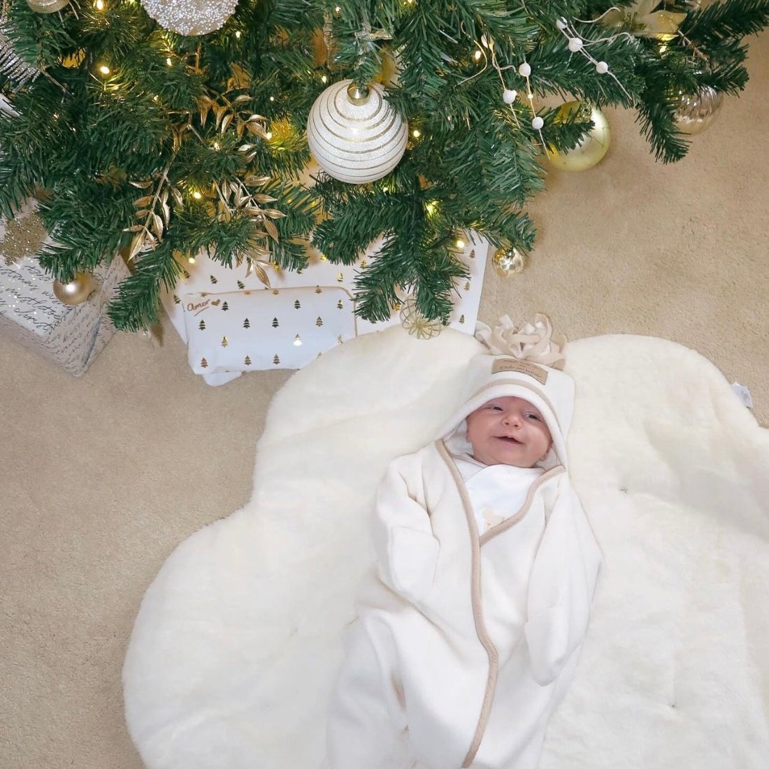 Newborn baby smiling snuggled up in the Cream Star Fleece Wrap Baby Blanket underneath the Christmas tree | Baby Blankets & Swaddles | Nursery Linen | Baby Christmas Gift Ideas - Clair de Lune UK