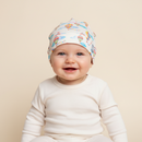 A smiling baby wearing a cream-colored long-sleeve outfit and the GrowGrows Up, Up and Away Baby Hat with a colourful hot air balloon pattern, set against a solid beige background. | Bamboo Babywear - Clair de Lune UK
