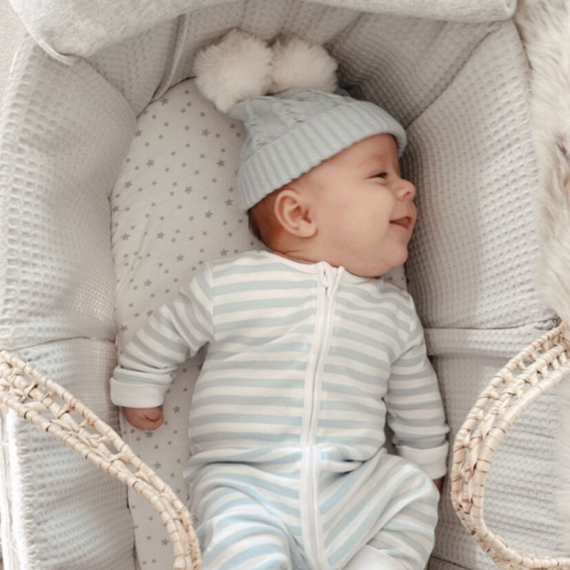 Cute baby wearing a striping jumpsuit and a cute pom pom hat is smiling inside Clair de Lune Waffle Palm Moses basket and Stars & Stripes Moses Basket Sheets