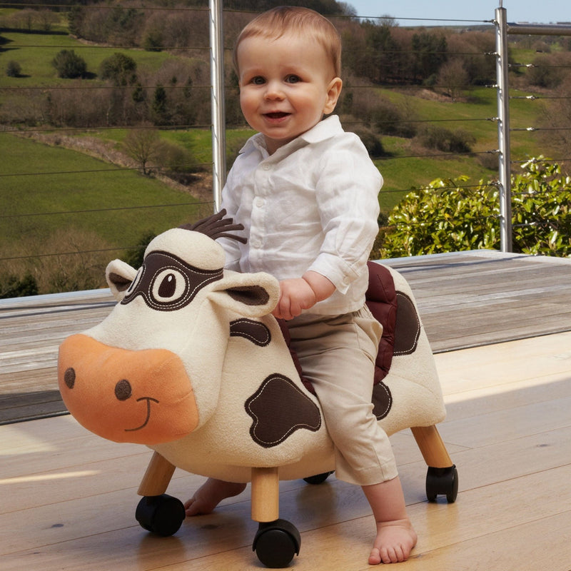 Toddler boy riding his favourite friend Little Bird Told Me Moobert Cow Ride On Toy on a farm next to a highland | Baby Walkers and Ride On Toys | Montessori Activities For Babies & Kids | Toys | Baby Shower, Birthday & Christmas Gifts - Clair de Lune UK